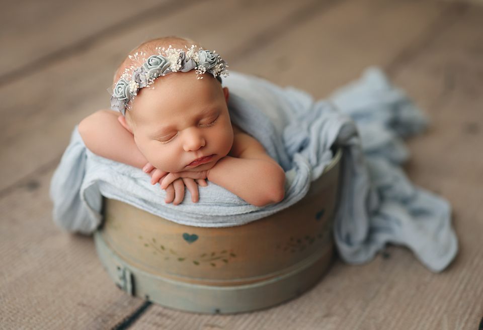 Sleeping newborn baby girl in a floral headband, nestled in a vintage bucket with a soft blue wrap on a wooden floor. Richmond newborn photo