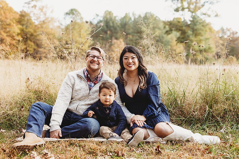 A smiling family of three sits together in a golden autumn meadow for a fall portrait Richmond VA family photography. Midlothian, Petersburg