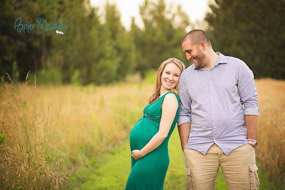 Pregnant couple smiling together in a sunny meadow, woman in green maternity dress holding her belly.