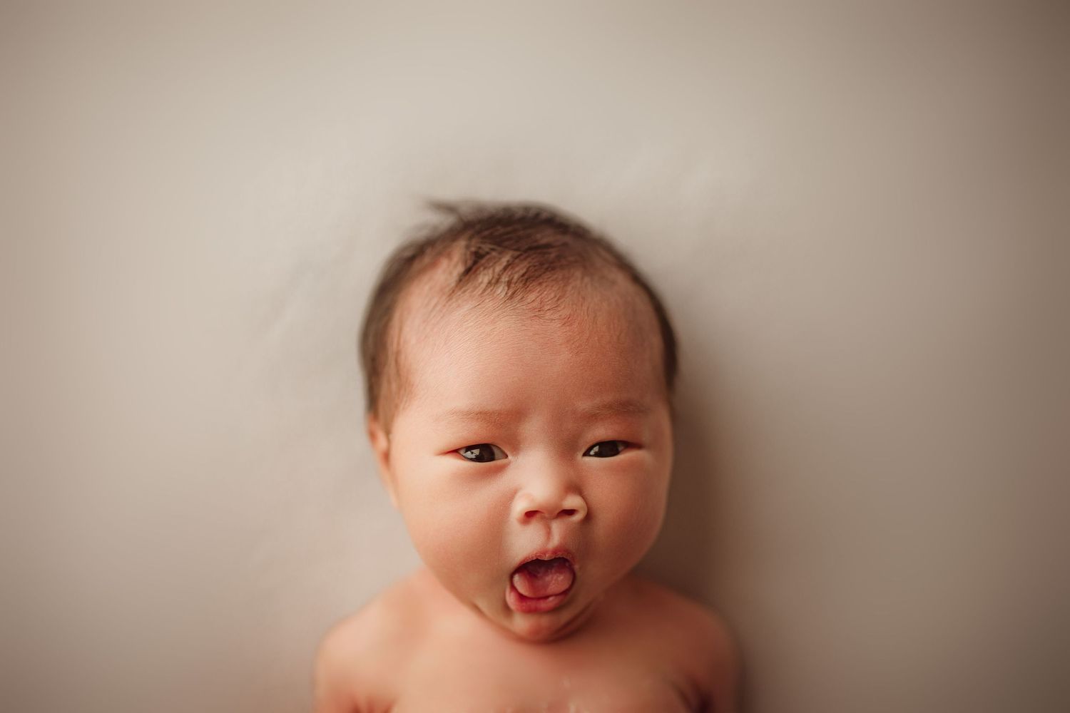 Newborn baby with dark hair lying on white background, mouth open in a yawn, eyes partially open.