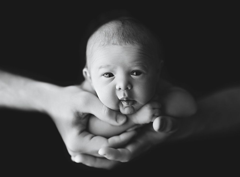 Black and white portrait of a newborn baby resting peacefully in adult hands against dark background.