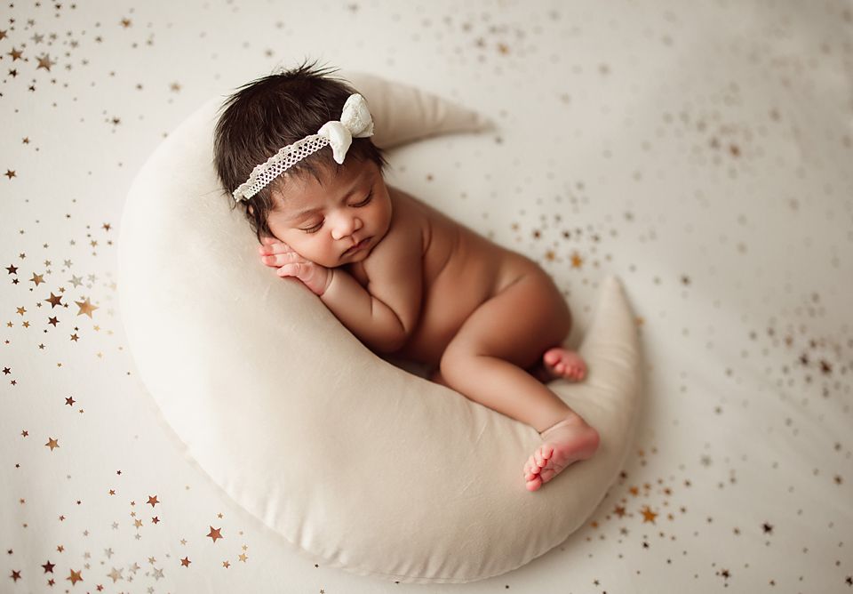Newborn baby sleeping peacefully on a crescent moon pillow surrounded by gold stars.