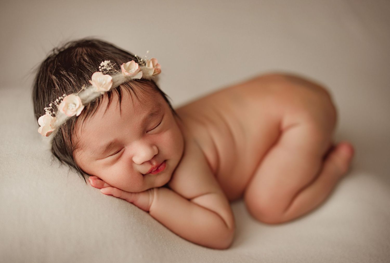Peaceful newborn baby girl sleeping with floral crown headband, posed on soft white blanket in studio portrait.