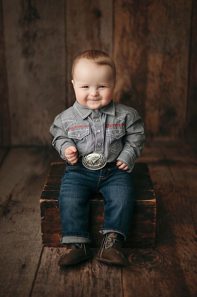 Smiling baby boy in cowboy shirt, jeans, and boots sitting on wooden crate for studio portrait.