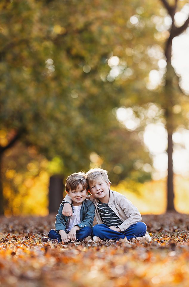 Two young boys sitting together in fallen autumn leaves, smiling in a sunny park. Richmond mini sessions photography