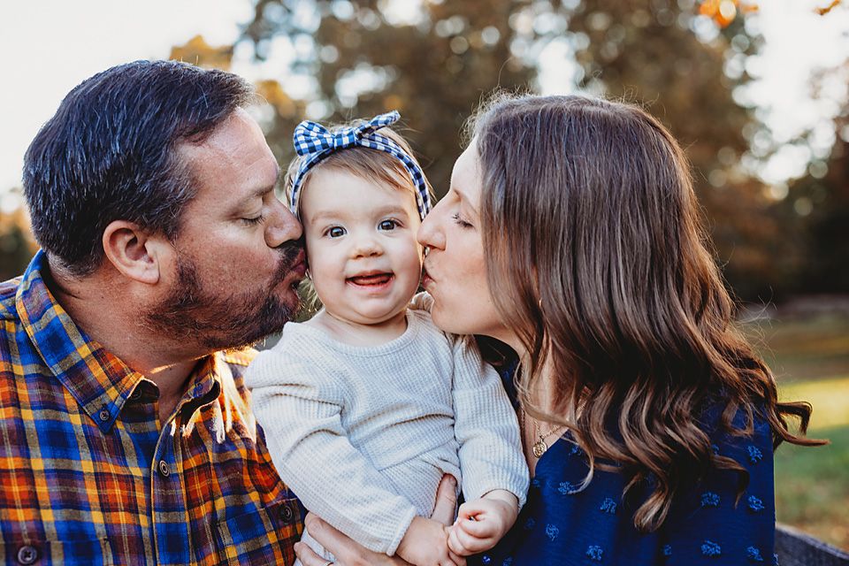 Parents kissing their smiling baby girl on the cheeks during an outdoor fall family portrait session.
