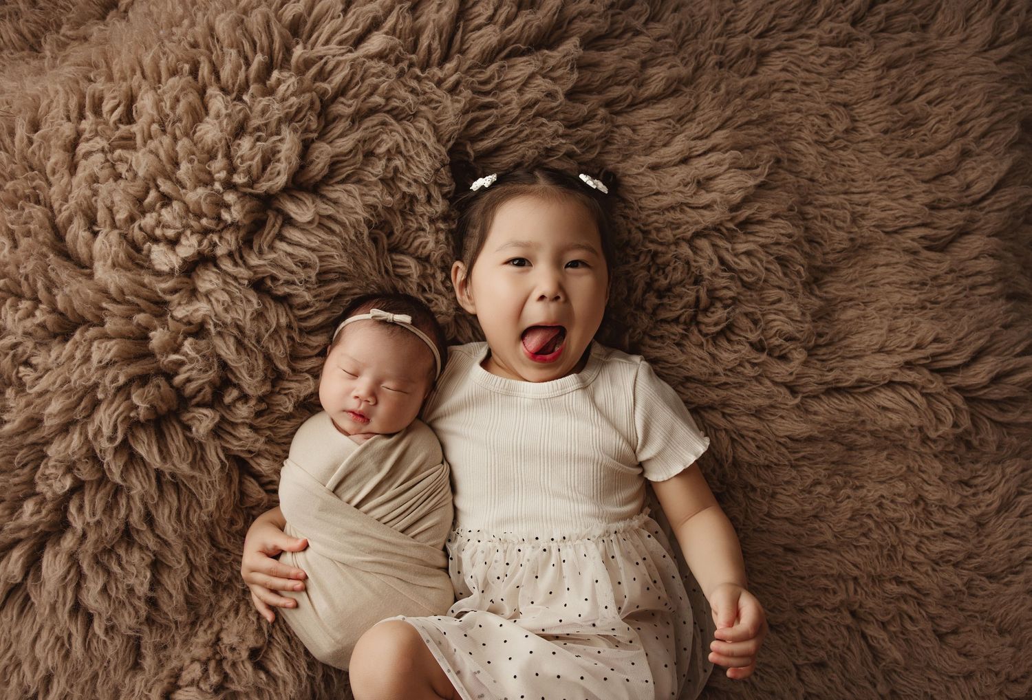 Two young sisters lying on a fluffy brown rug, newborn swaddled beside excited toddler with bow headband.