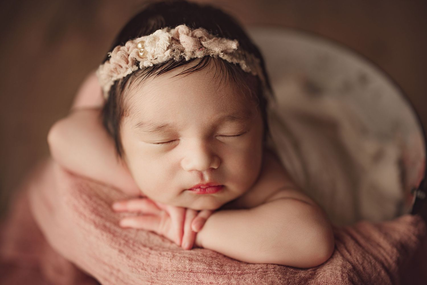 Peaceful newborn baby girl sleeping in a bucket wrapped in pink, wearing a delicate floral headband.