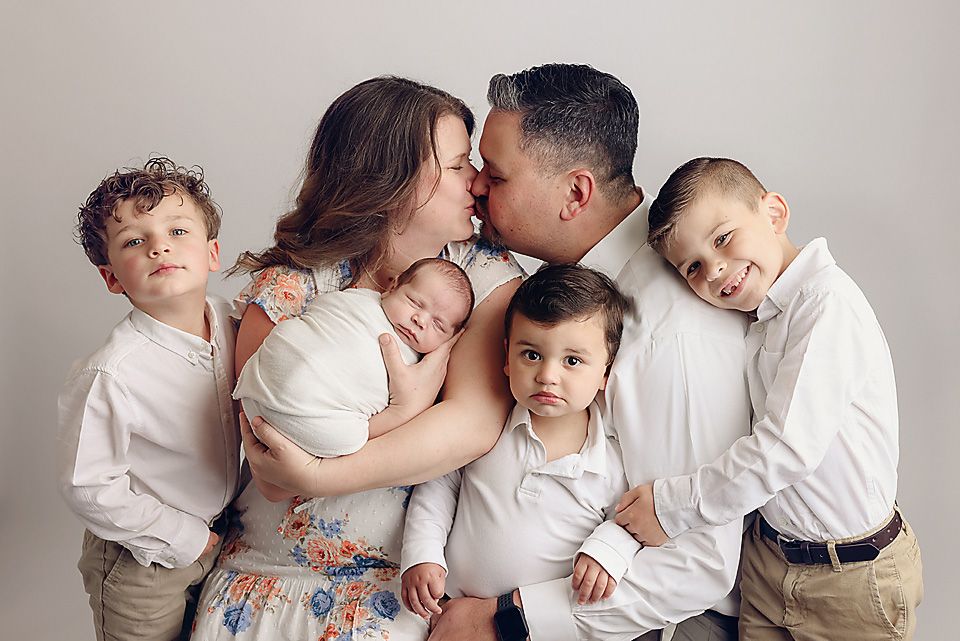 Family of six in white outfits with parents kissing while holding their newborn baby. Chesterfield va studio newborn photography session