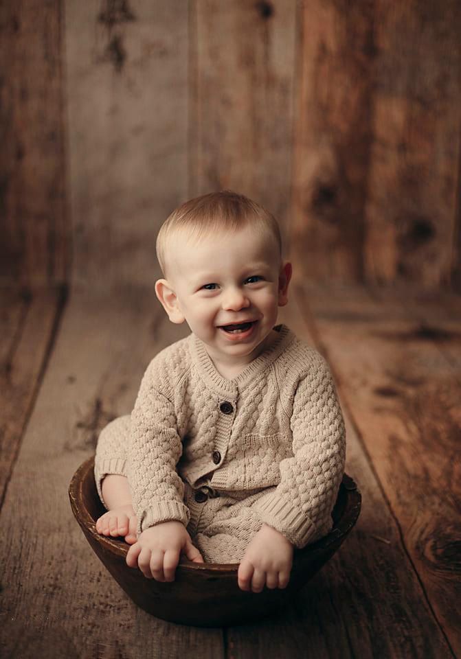 Happy laughing baby boy in knit romper sitting in wooden bowl against rustic wood backdrop.