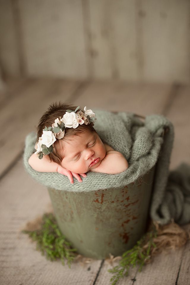 Sleeping newborn baby girl in a green bucket with a floral crown headband on a rustic wood floor.