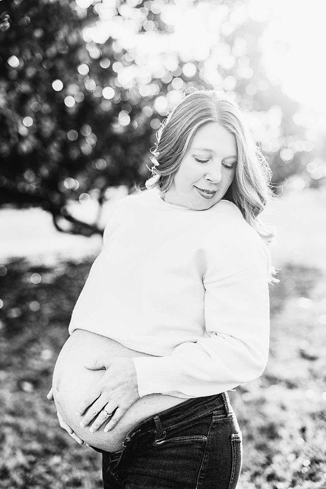 Black and white portrait of a pregnant woman cradling her baby bump outdoors in soft sunlight.