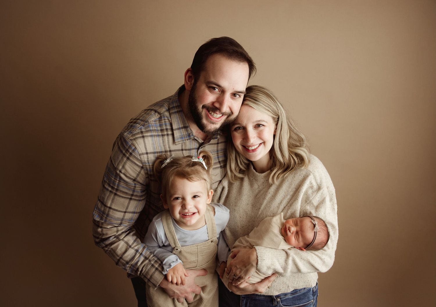 Happy family of four posing together, parents with toddler and newborn baby against beige background.