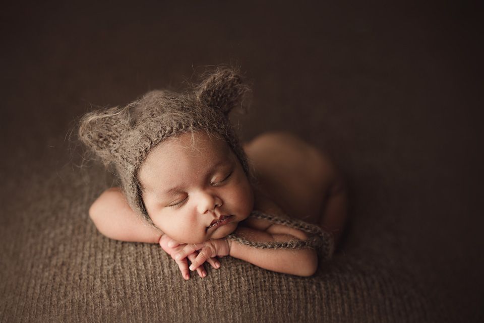 Sleeping newborn baby wearing a knit bear ear bonnet posed on brown textured backdrop.