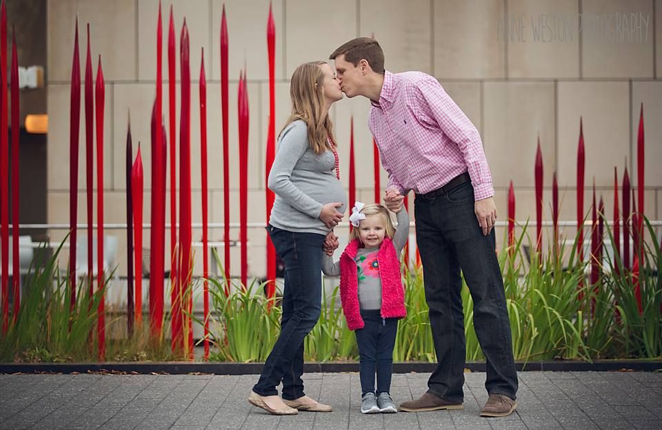 A young family with a toddler girl kisses in front of red glass sculptures outdoors. Richmond, VA maternity photographer.