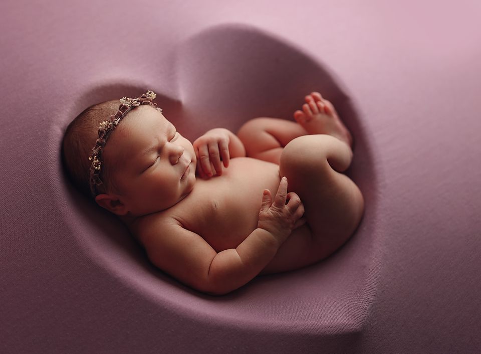 Sleeping newborn baby girl with floral headband curled inside a pink heart-shaped prop.