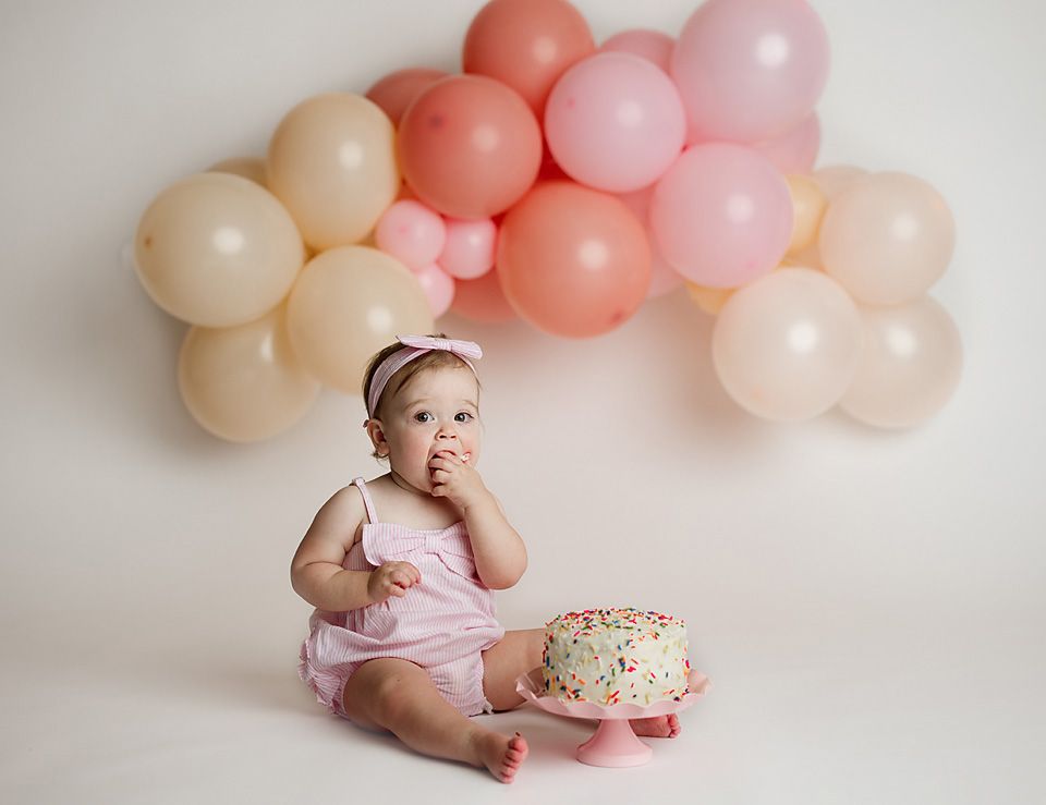 Baby girl in pink romper sitting beside a smash cake surrounded by pastel balloon garland.