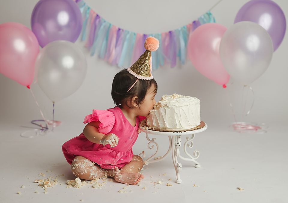 Baby girl in pink dress smashing face into white first birthday cake surrounded by colorful balloons. cake smash Richmond, Chesterfield