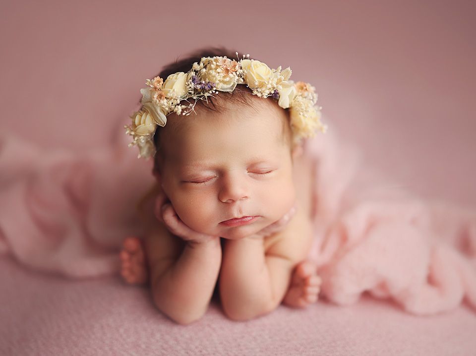 Newborn baby girl sleeping peacefully wearing a delicate floral crown on a soft pink backdrop.