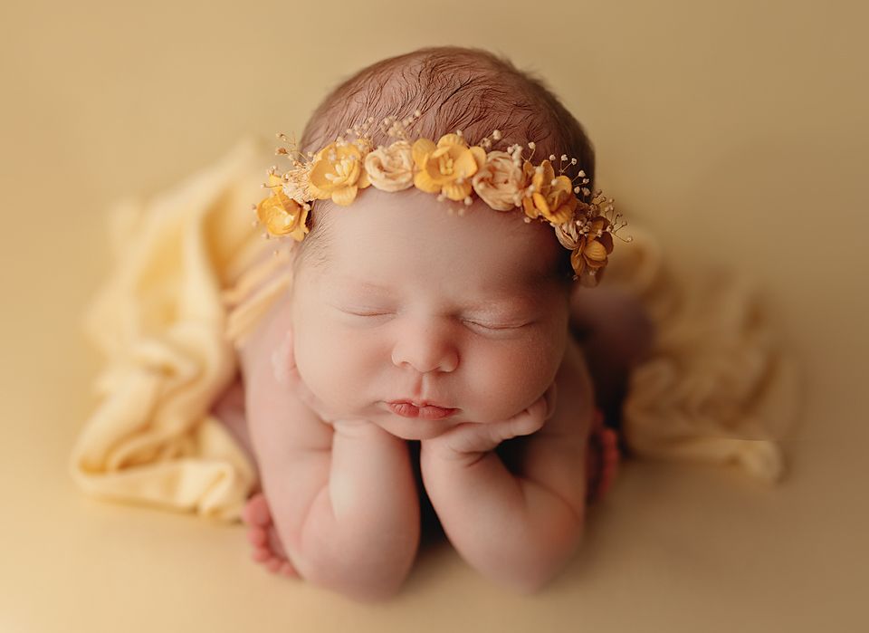 Sleeping newborn baby wearing a yellow floral headband, posed with hands under chin on a soft golden yellow blanket. froggy pose