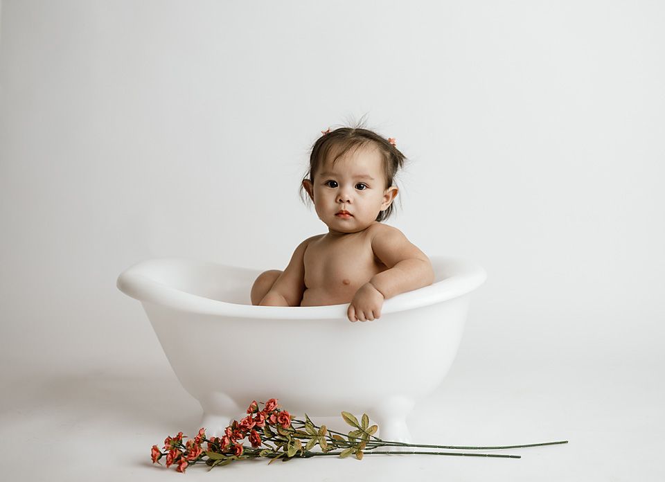 Cute baby sitting in a white mini clawfoot bathtub with red flowers on a white background.