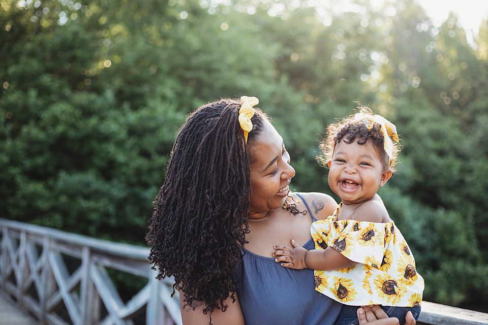 Joyful mother holding laughing baby girl in matching yellow sunflower outfits outdoors.
