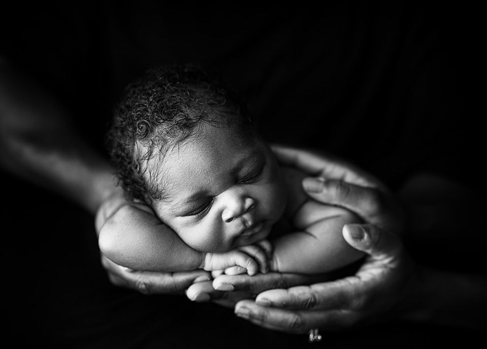 Black and white close-up of newborn baby sleeping peacefully in loving hands against dark background.