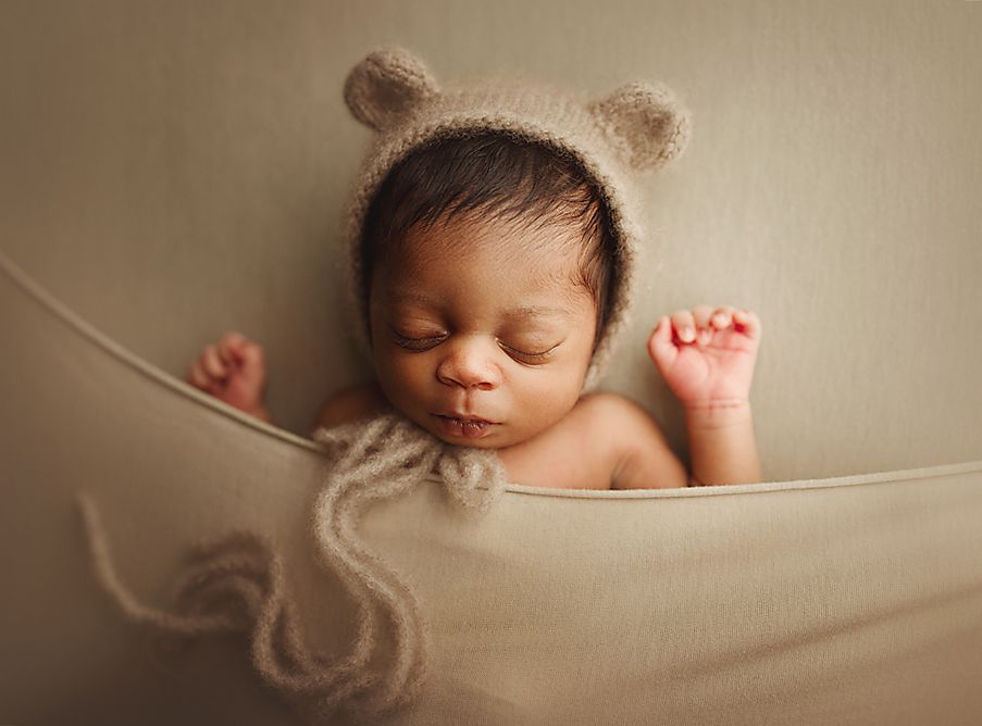 Newborn baby sleeping in a cozy fabric hammock wearing an adorable knit bear ear bonnet.