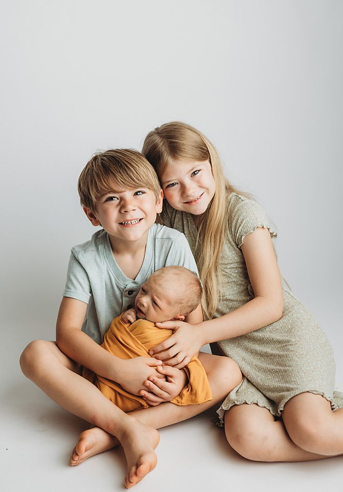 Two smiling siblings sitting together on white floor proudly holding their newborn baby brother newborn photographer Richmond Chesterfield