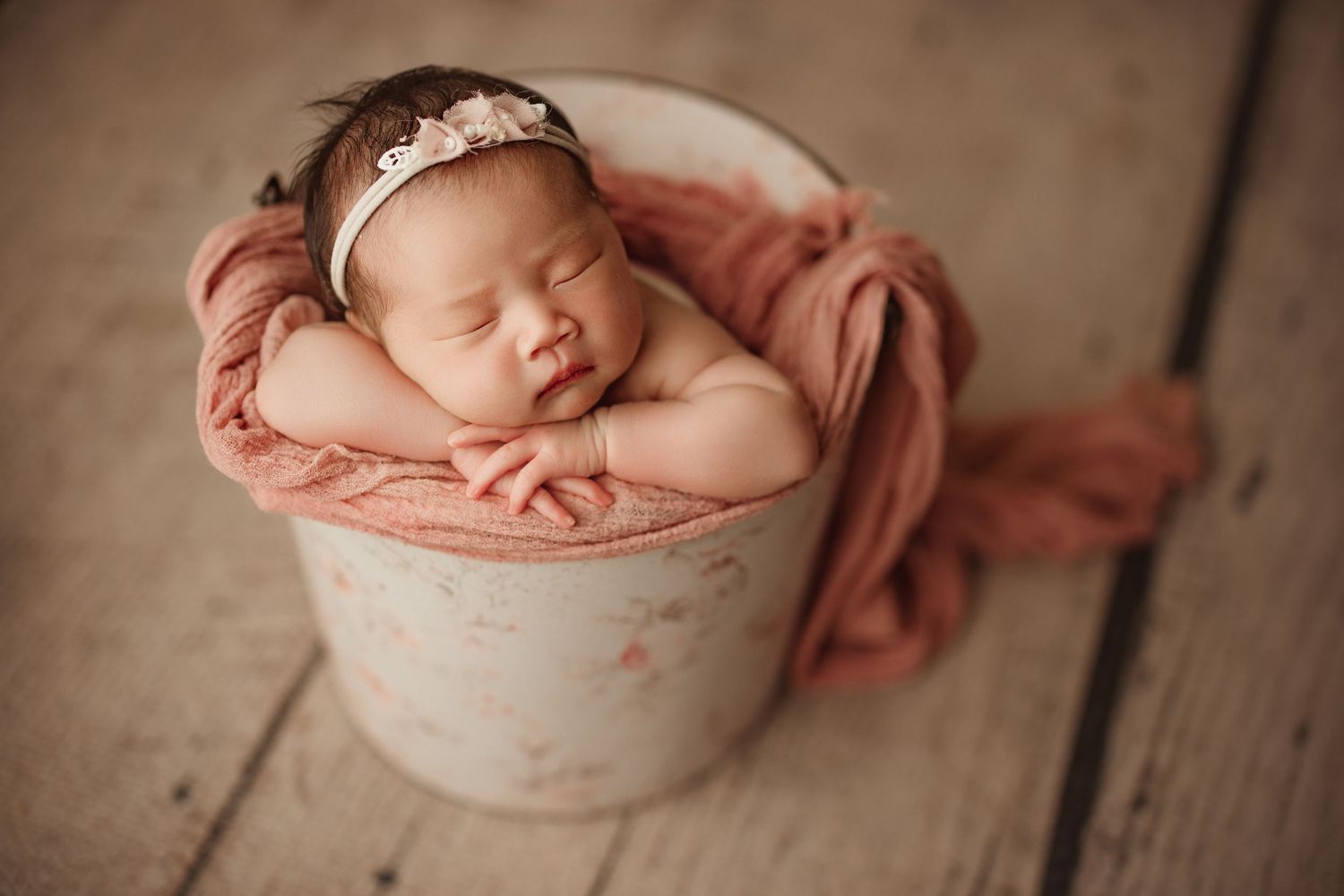 Newborn baby girl sleeping peacefully in a white bucket with pink wrap and floral headband on wood floor. Richmond newborn photographer