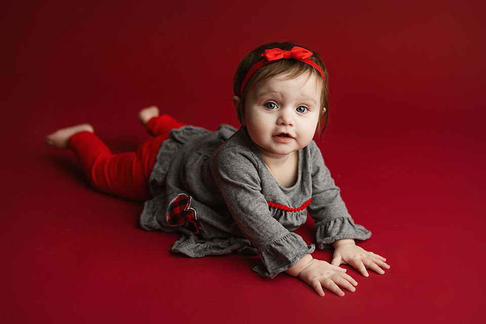 Baby girl in red and gray outfit lying on a red background with a red bow headband.