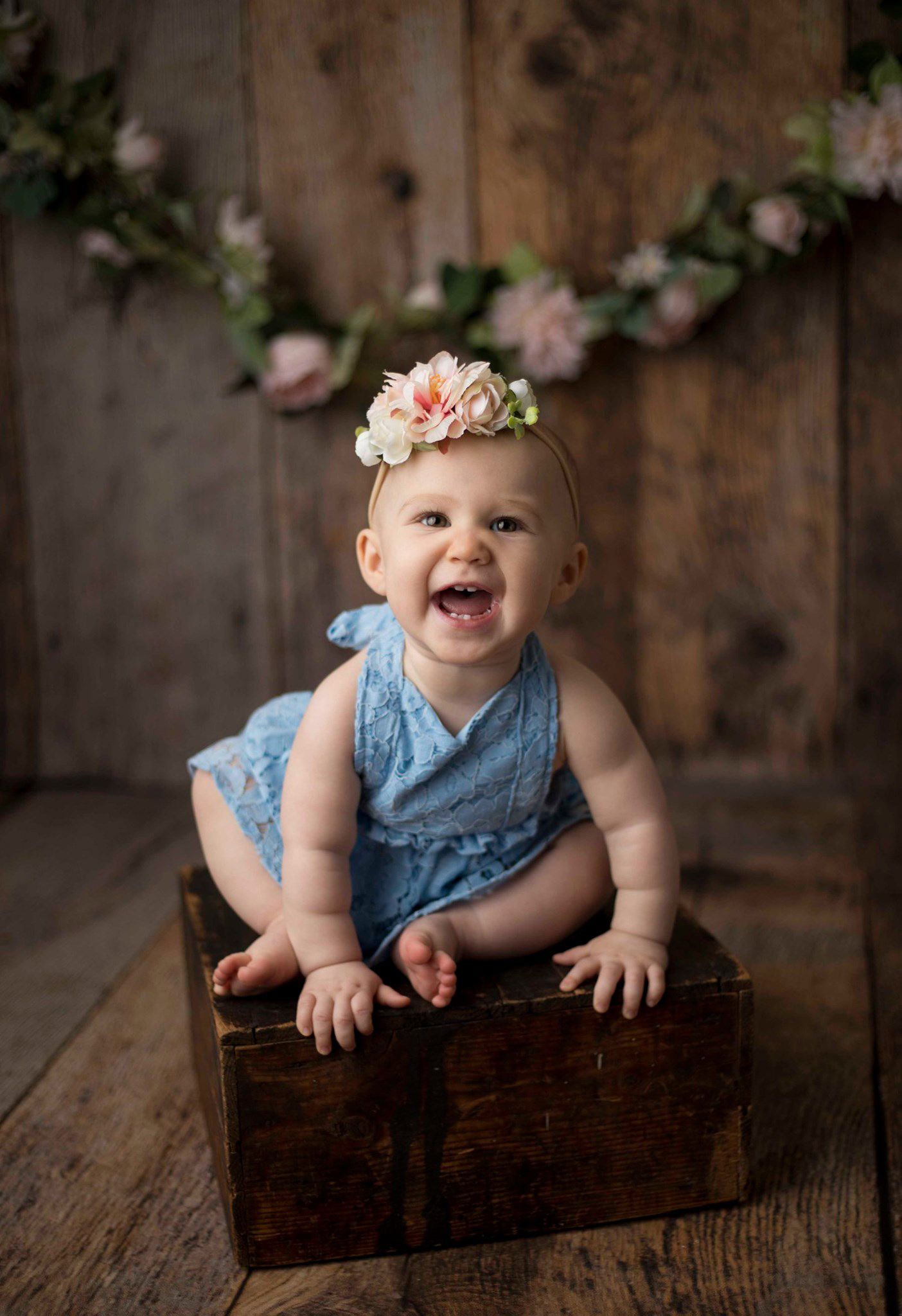 Laughing baby girl in blue romper and floral crown sitting in a rustic wooden crate. Milestone 1 year photography session Chesterfield.
