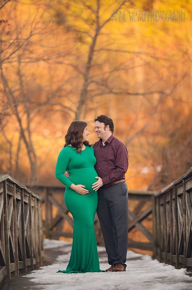 Pregnant woman in green gown with partner on snowy bridge, fall foliage backdrop, maternity photography session. Richmond VA Petersburg,