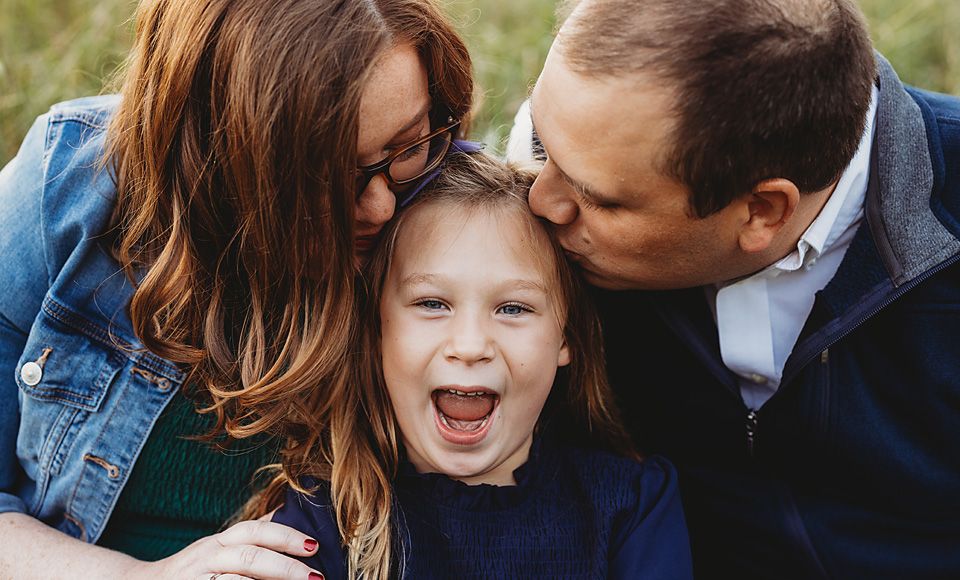 Laughing child being kissed on both cheeks by her smiling parents outdoors.