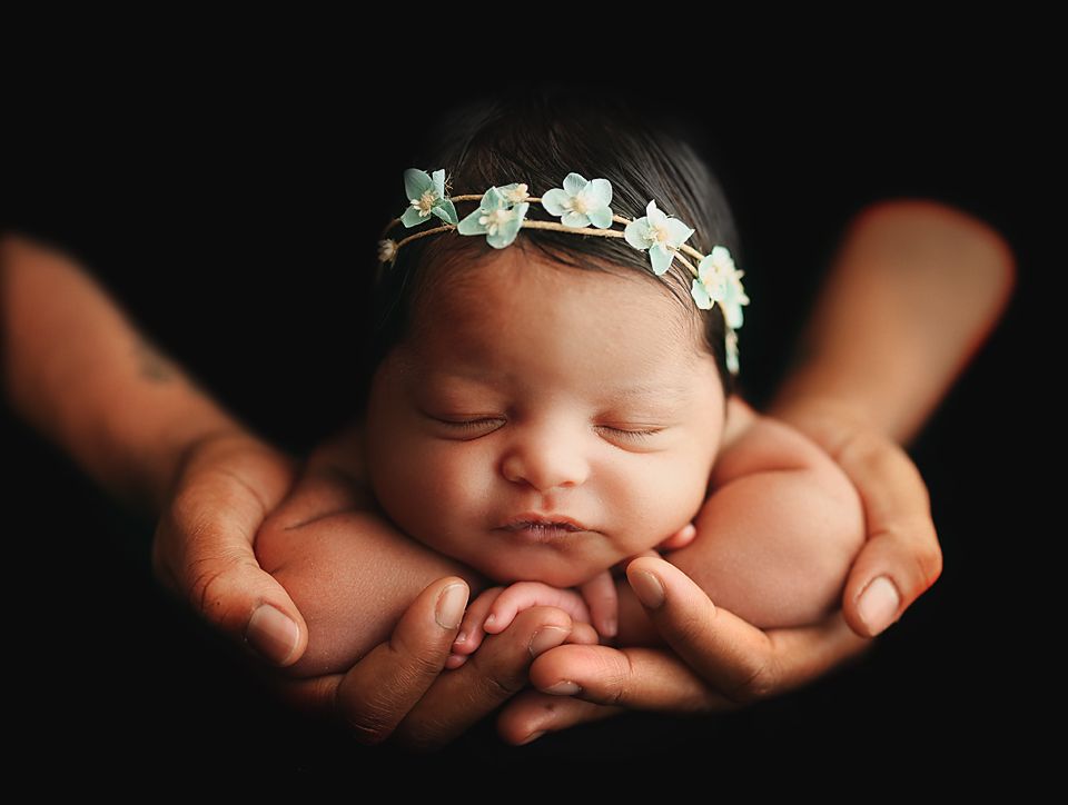 Newborn baby girl with floral headband sleeping peacefully in cupped hands against dark background.