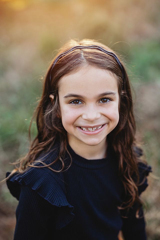 Smiling young girl with brown hair wearing navy dress in sunny outdoor setting.