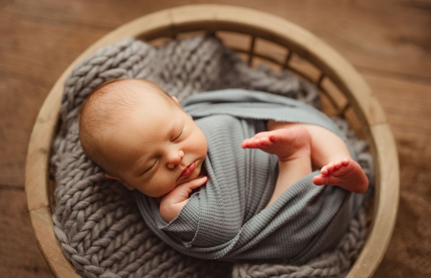 Newborn baby swaddled in gray wrap sleeping peacefully in a round wicker basket with knit blanket. Richmond newborn photographer