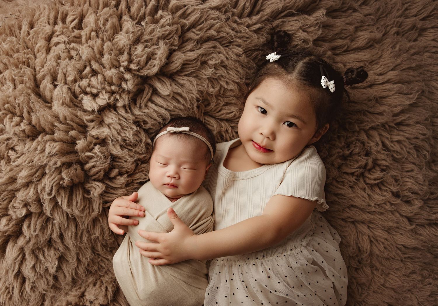 Young girl with hair clips gently hugs sleeping newborn baby wrapped in beige on a fluffy brown rug.