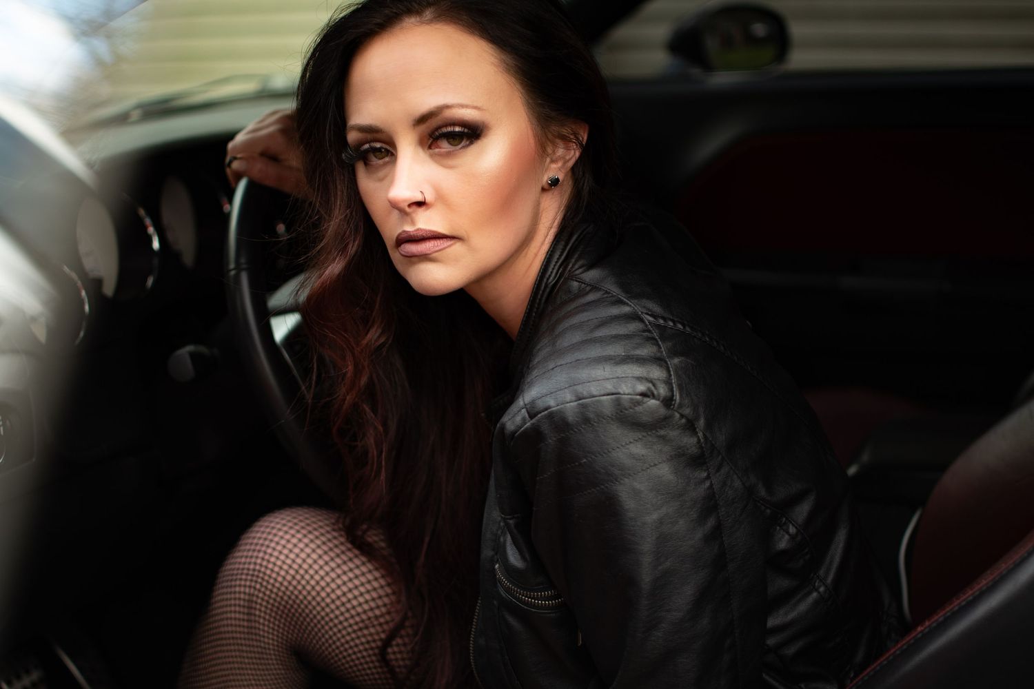 Portrait of a woman in black leather jacket posing in car with dramatic lighting.