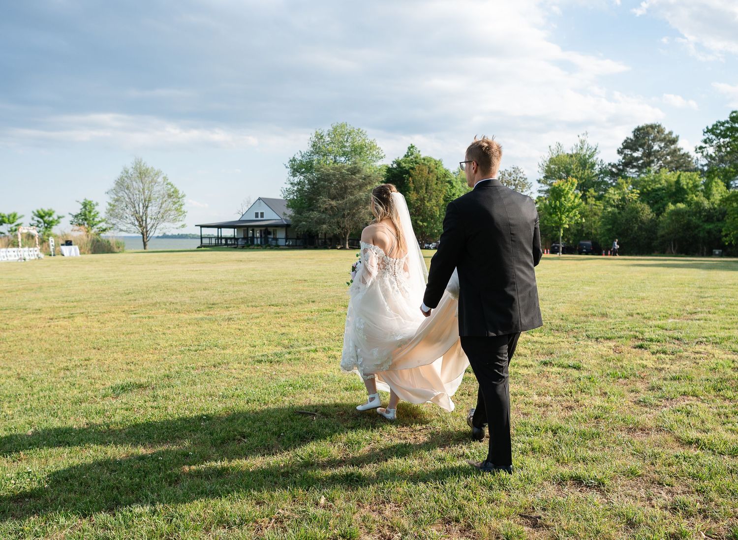 Bride and groom walking hand in hand across a green lawn toward a white venue building on a sunny wedding day.