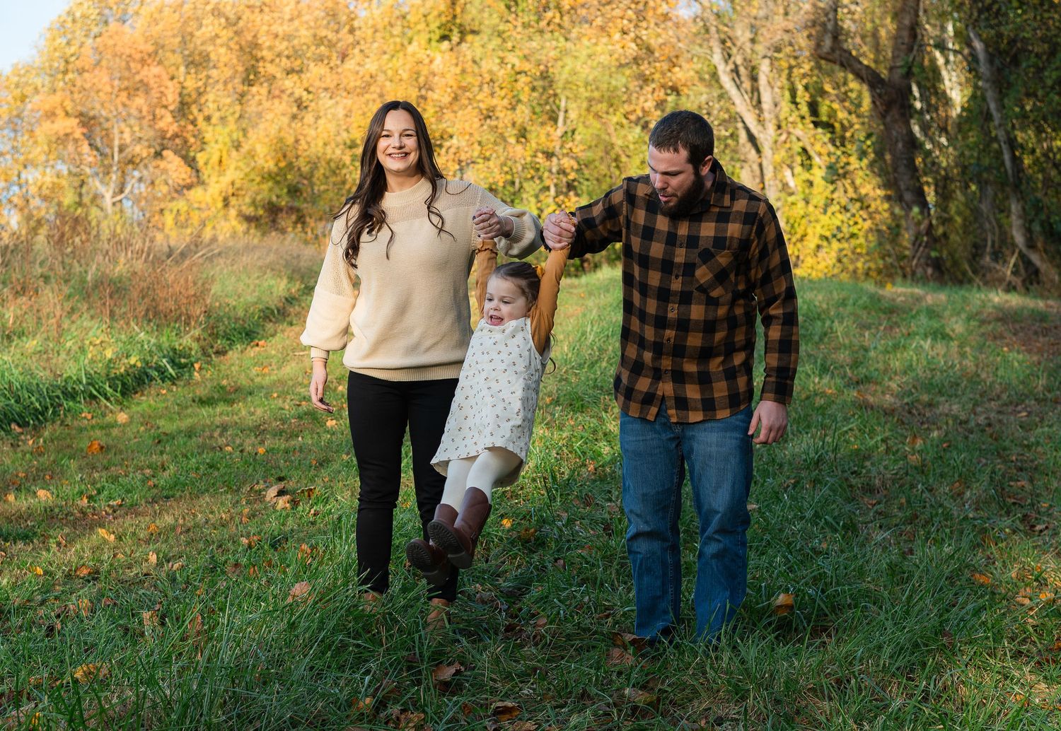 Family enjoys an autumn walk together while swinging child between them on a path surrounded by golden fall foliage.
