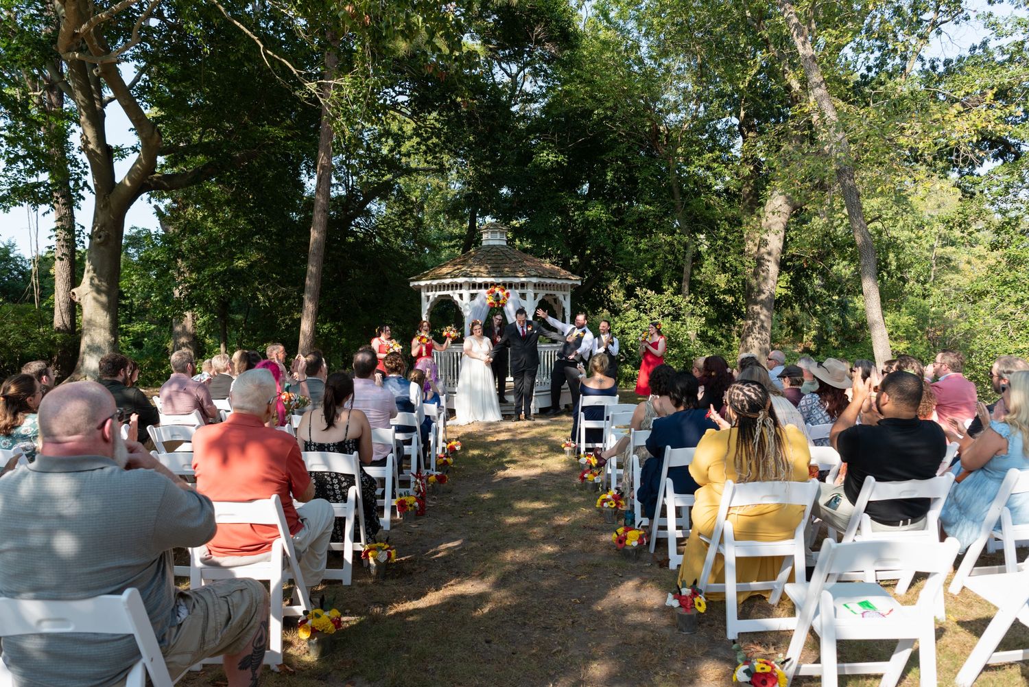 Outdoor wedding ceremony with white chairs and gazebo surrounded by trees.