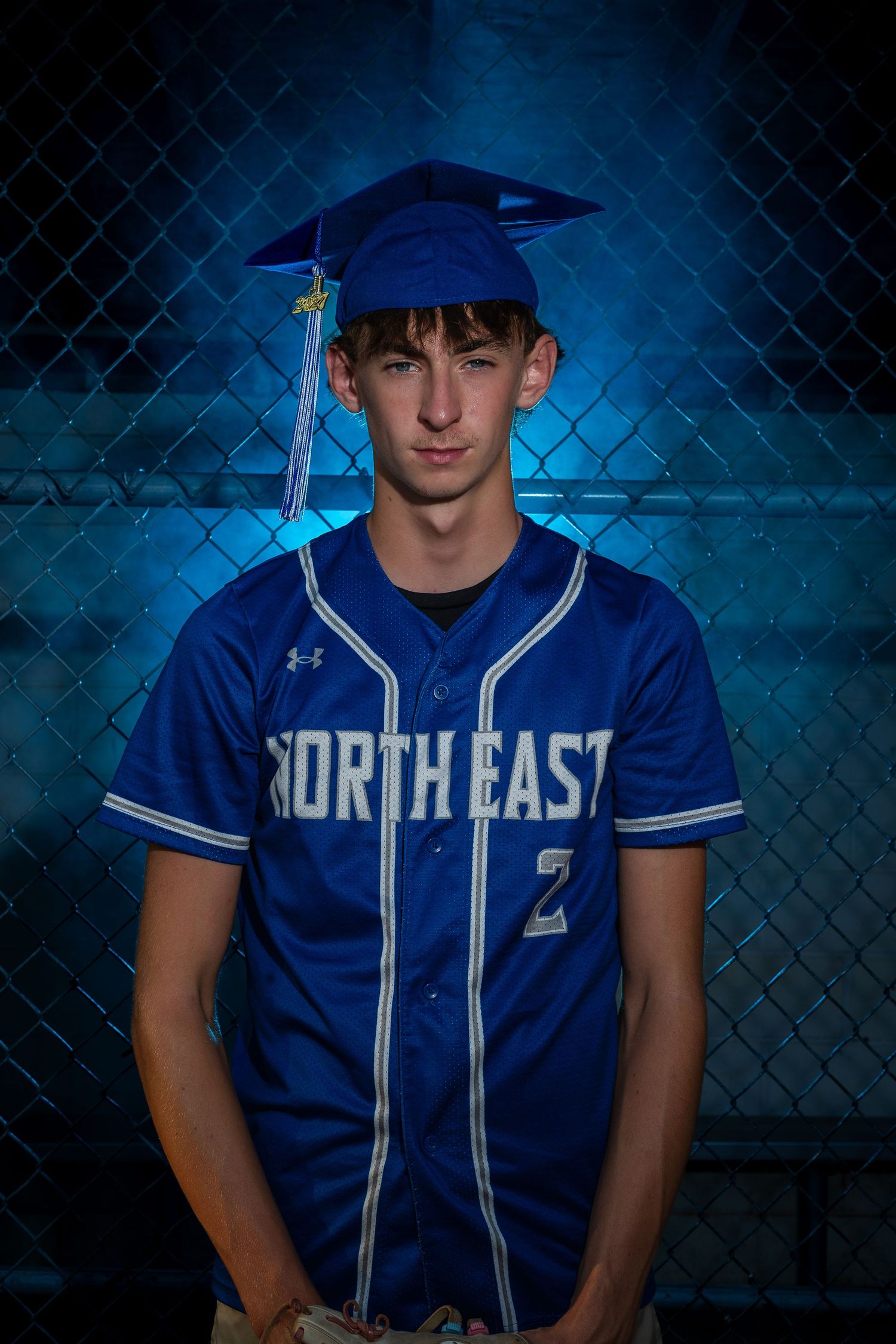 Young man in Northeast baseball jersey #2 and blue graduation cap poses by chain-link fence.