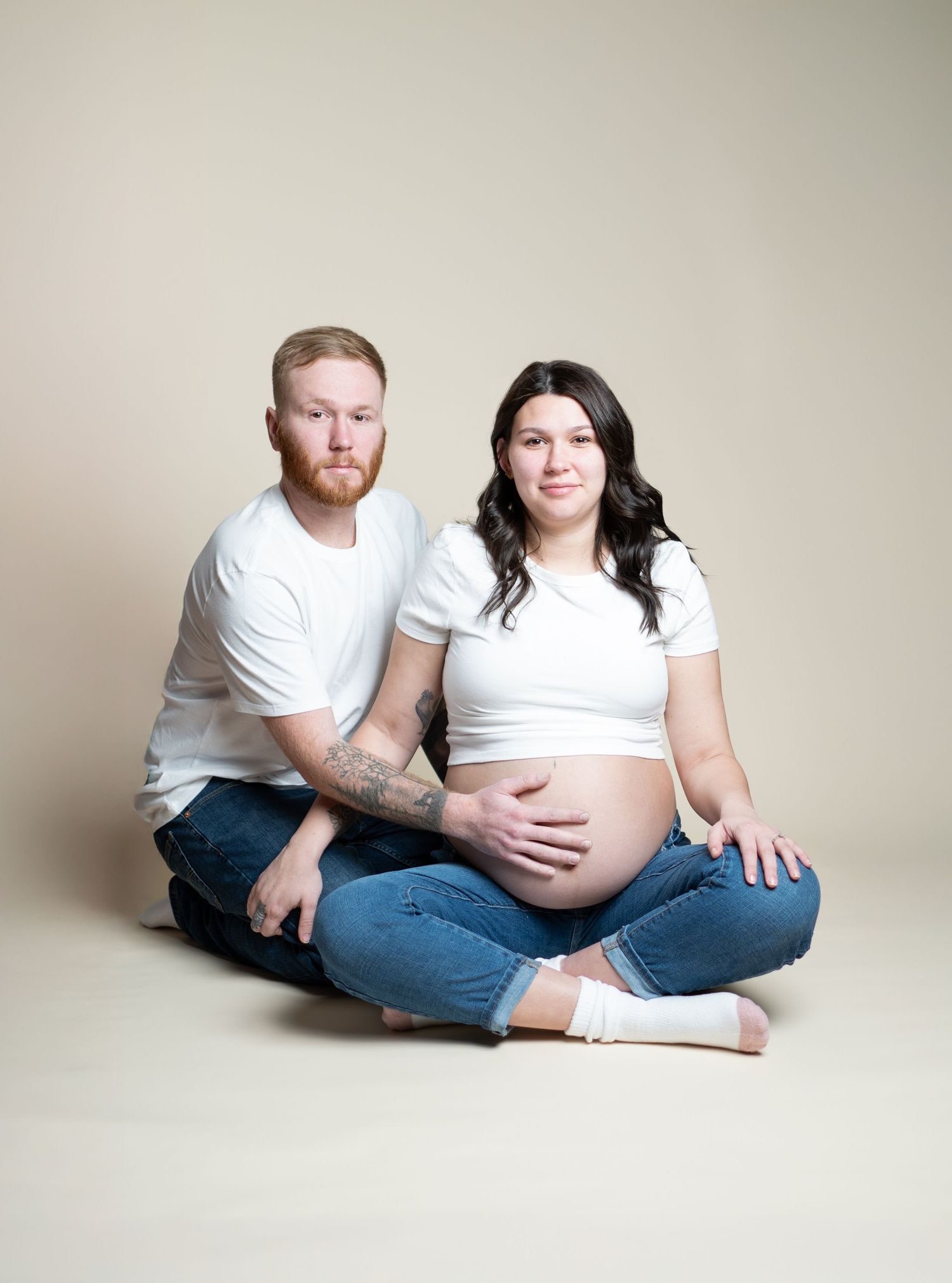 Couple in white shirts and jeans sitting on beige backdrop during intimate maternity photoshoot.