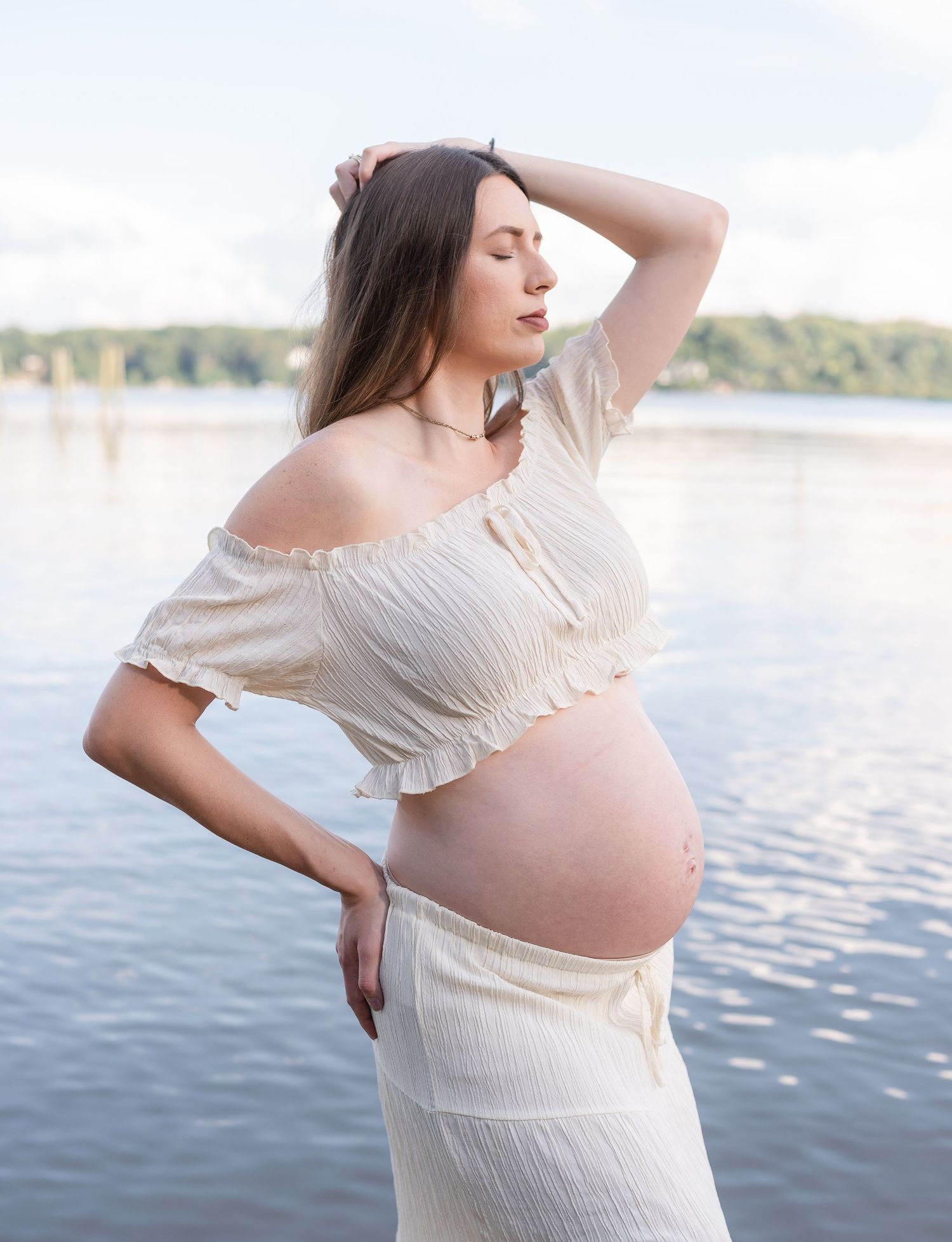 Pregnant woman in white crop top and skirt poses by a lake, hand in hair, showing baby bump outdoors.