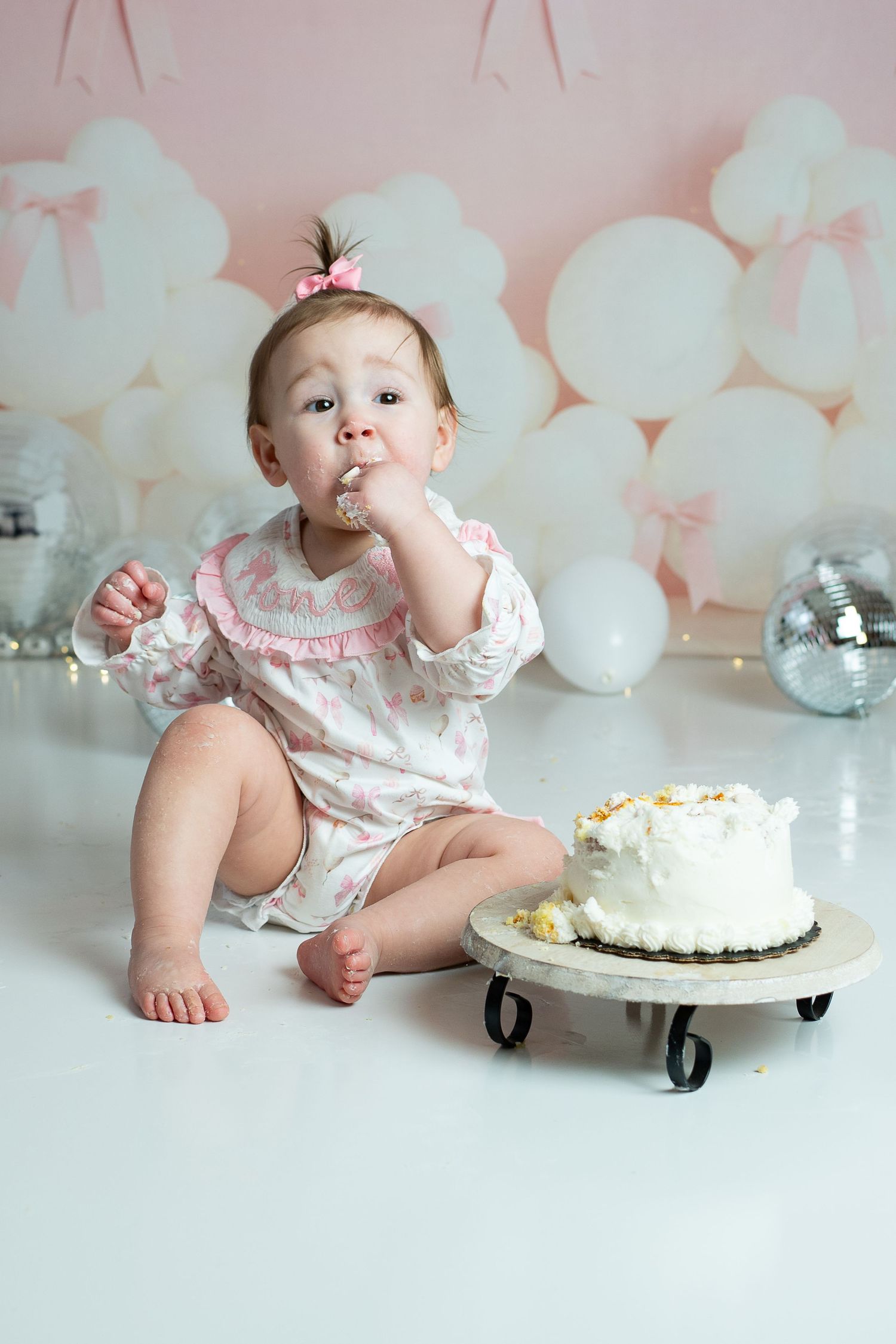 A baby in a floral dress enjoys a cake smash photo session with white balloons and silver disco balls in the background.
