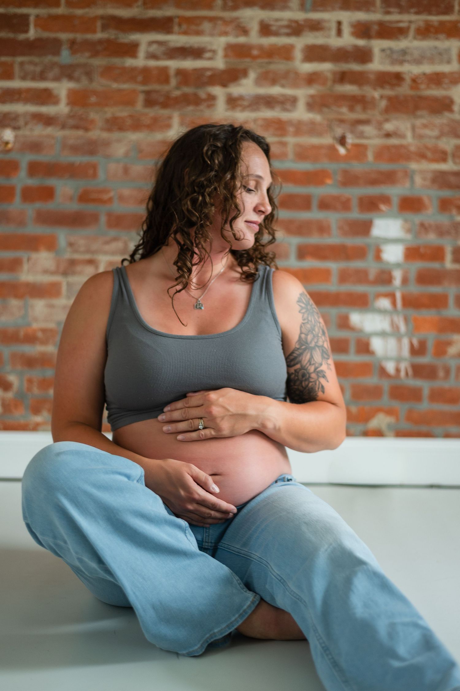 Pregnant woman with tattoo sitting cross-legged on floor, cradling bare belly, wearing gray tank and jeans against brick wall.