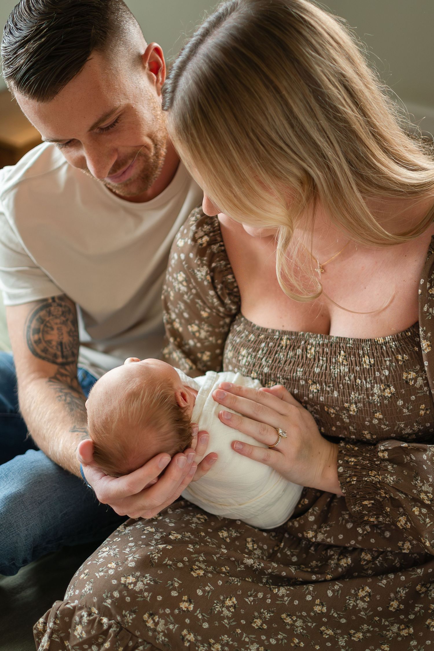 A couple in casual attire shares tender moments with their newborn baby while sitting on a brown couch.