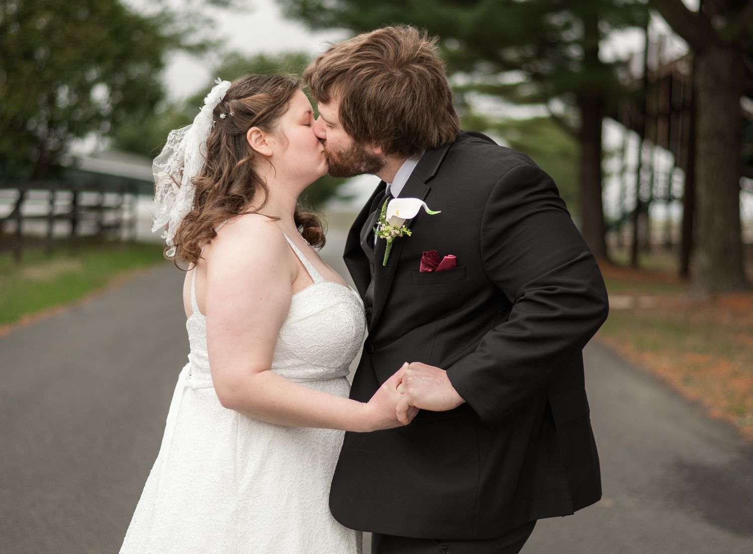 Newlywed couple shares a romantic kiss on a tree-lined path in their wedding attire.
