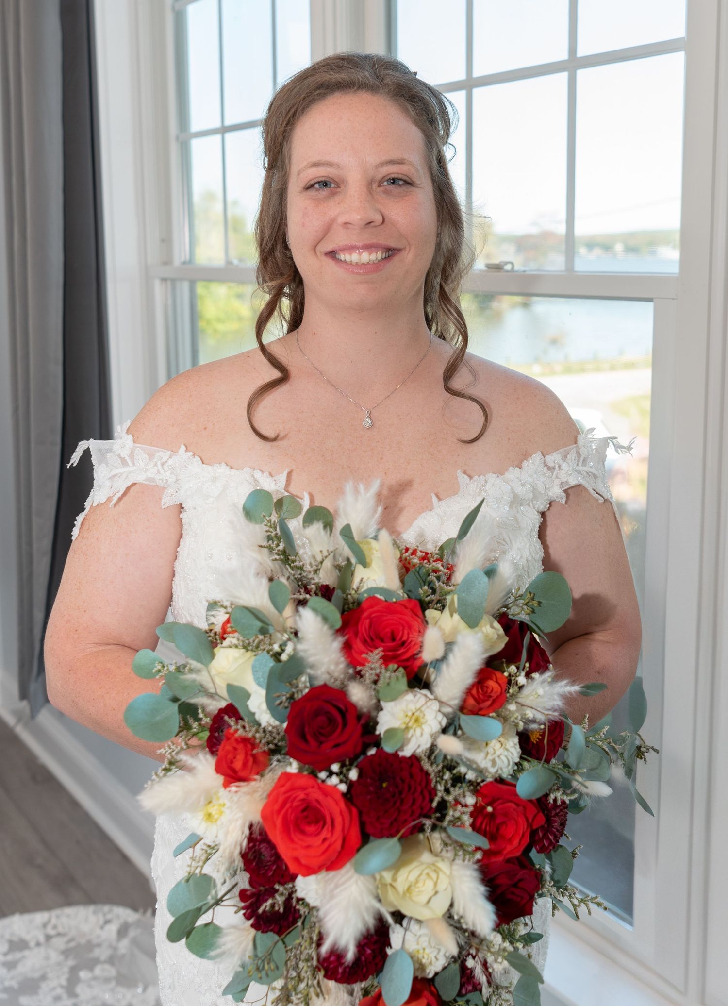 Bride holding red and white bouquet poses by bright window in off-shoulder lace gown.